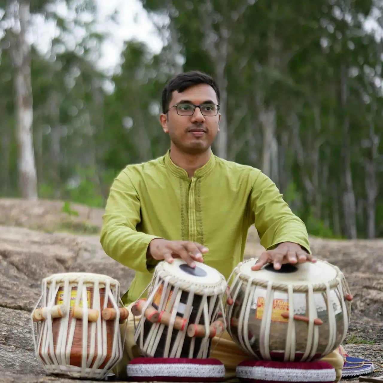 Kaushik Bhat playing tabla outdoors on a rock amid trees